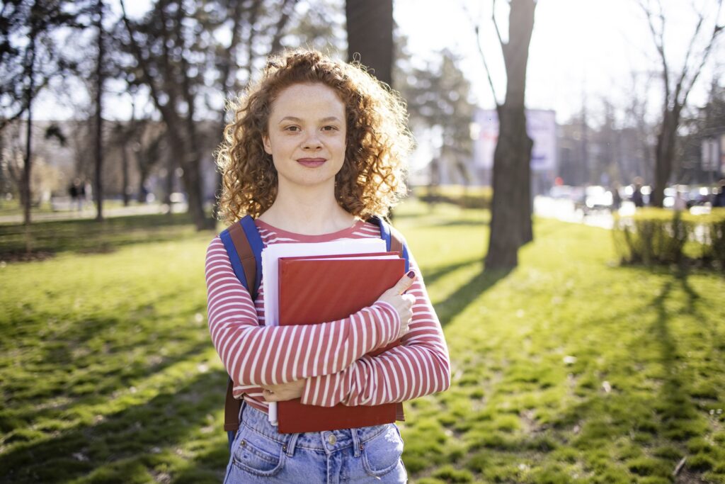 Portrait of a happy female university student with a red curly hair standing in a public park, holding her books and smiling